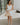 Image shows full body front view of female model wearing a white mini dress and holding a beige purse.