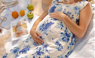 image shows blonde female model laying down on blanket while holding pregnant belly. Model is wearing white dress with blue floral print.
