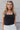 Front view of model wearing the Luna Black Sleeveless Bodysuit which features black knit fabric, a square neckline with a racerback, a sleeveless body, and a thong bottom with snap closures.
