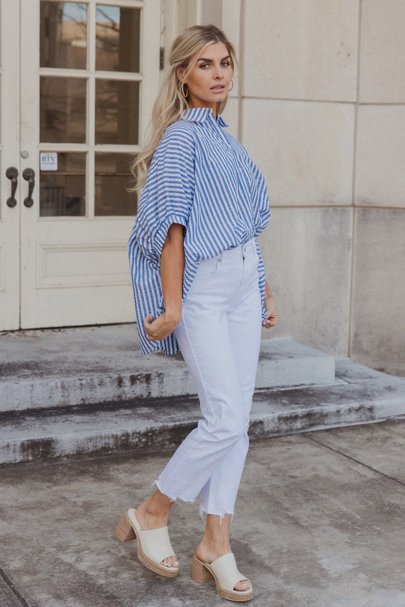 Full body view of model wearing the Ella Blue & White Stripe Button Up Top which features dark blue and white fabric with a striped pattern, white button-up front closures, an oversized fit, a collared neckline and short sleeves with cuffs.