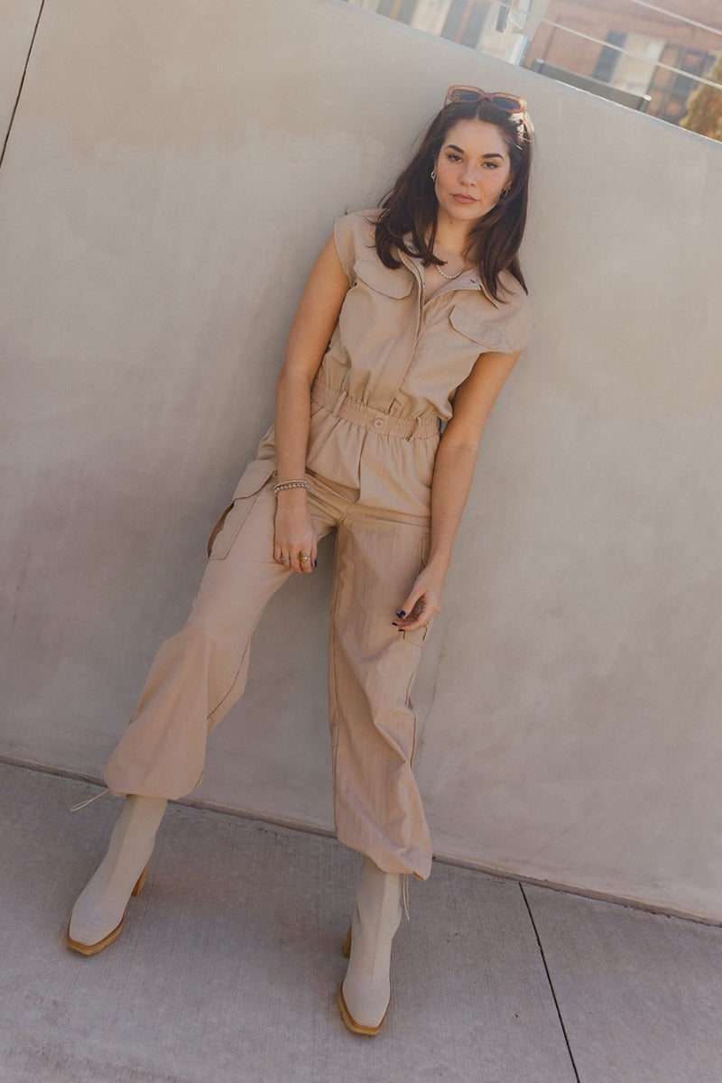 Full body view of model wearing the Zoe Khaki Sleeveless Cargo Jumpsuit which features washed denim fabric, mini length, a small slit detail, a monochrome side zipper with a hook closure, and a monochrome adjustable belt with a buckle.