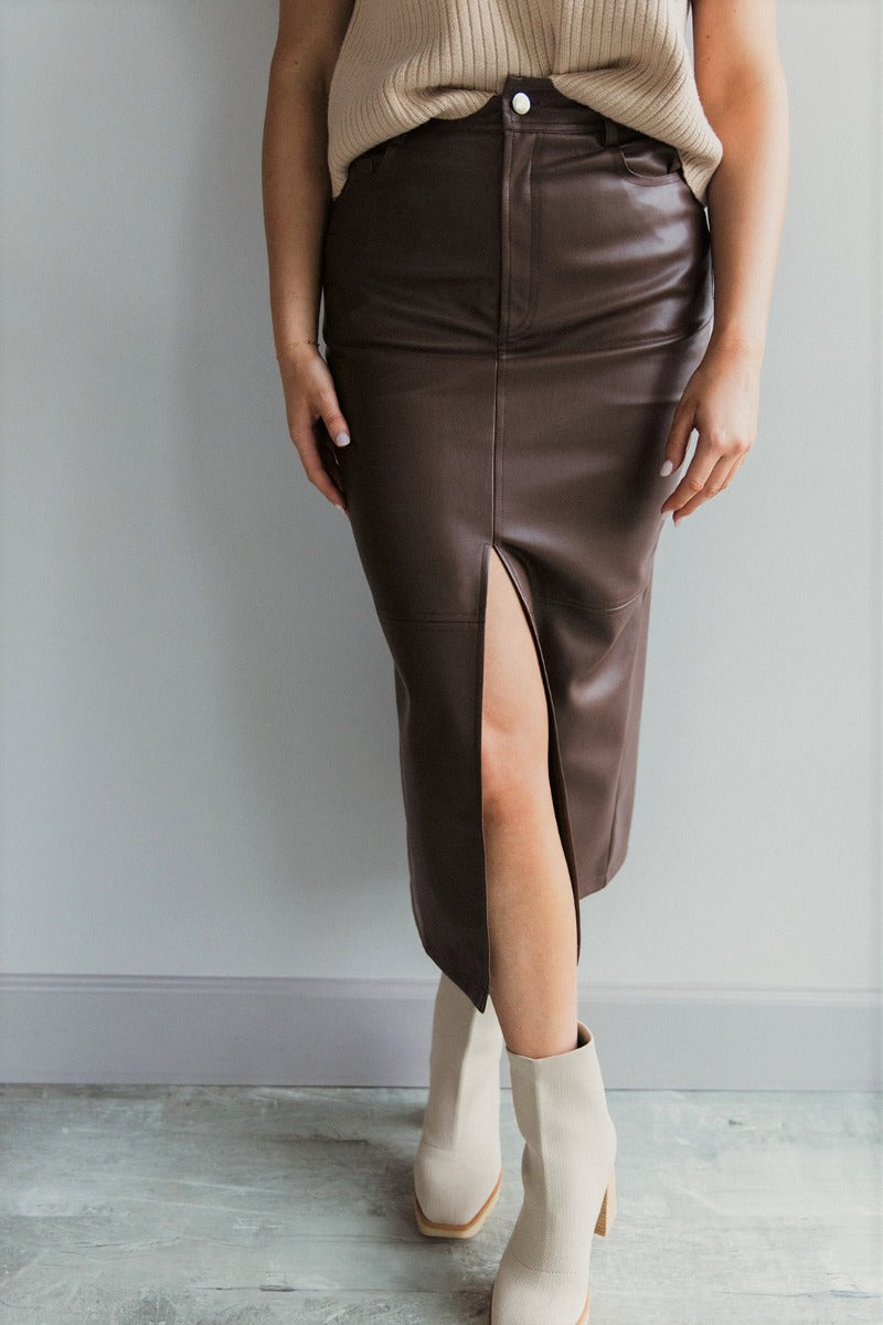 front view of model wearing the Trouble Maker Pleather Midi Skirt that has dark brown faux leather, midi length, a slit, pockets, a front zipper, and belt loops