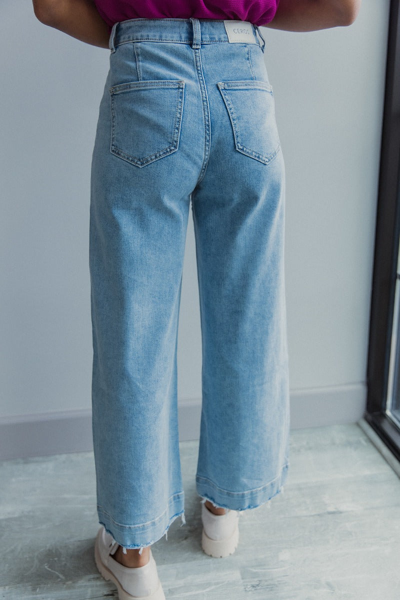 Back view of model wearing the Ceros: Here Not There Jeans which features light denim wash fabric, two front pockets, two back pockets, front zipper with button closure, belt loops, super high waisted, wide cropped flares and distressed hem.