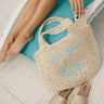 Image shows the Sea You Later Natural Tote that has natural woven material with blue lettering that says "sea you later". Model is sitting in pool chair holding the tote.