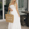Full body front view of model wearing the Paloma White Eyelet Strapless Maxi Dress that has a strapless upper with eyelet and floral details and a tiered maxi skirt. Model is holding "beach please" tote bag
