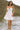 Full body view of female model wearing the Ayla Off White Key Hole Smocked Mini Dress which features Off White Lightweight Fabric, Ruffle Hem Skirt, Mini Length, Smocked Upper with Tie Details, Key Hole Designs, Adjustable Straps and Sleeveless