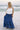 Full body front view of female model wearing the Madelyn White Tie Strap Tank Top that has tiered white fabric and tie straps with navy trim. Worn with blue skirt.