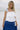 Upper body front view of female model wearing the Madelyn White Tie Strap Tank Top that has tiered white fabric and tie straps with navy trim. Worn with blue skirt.