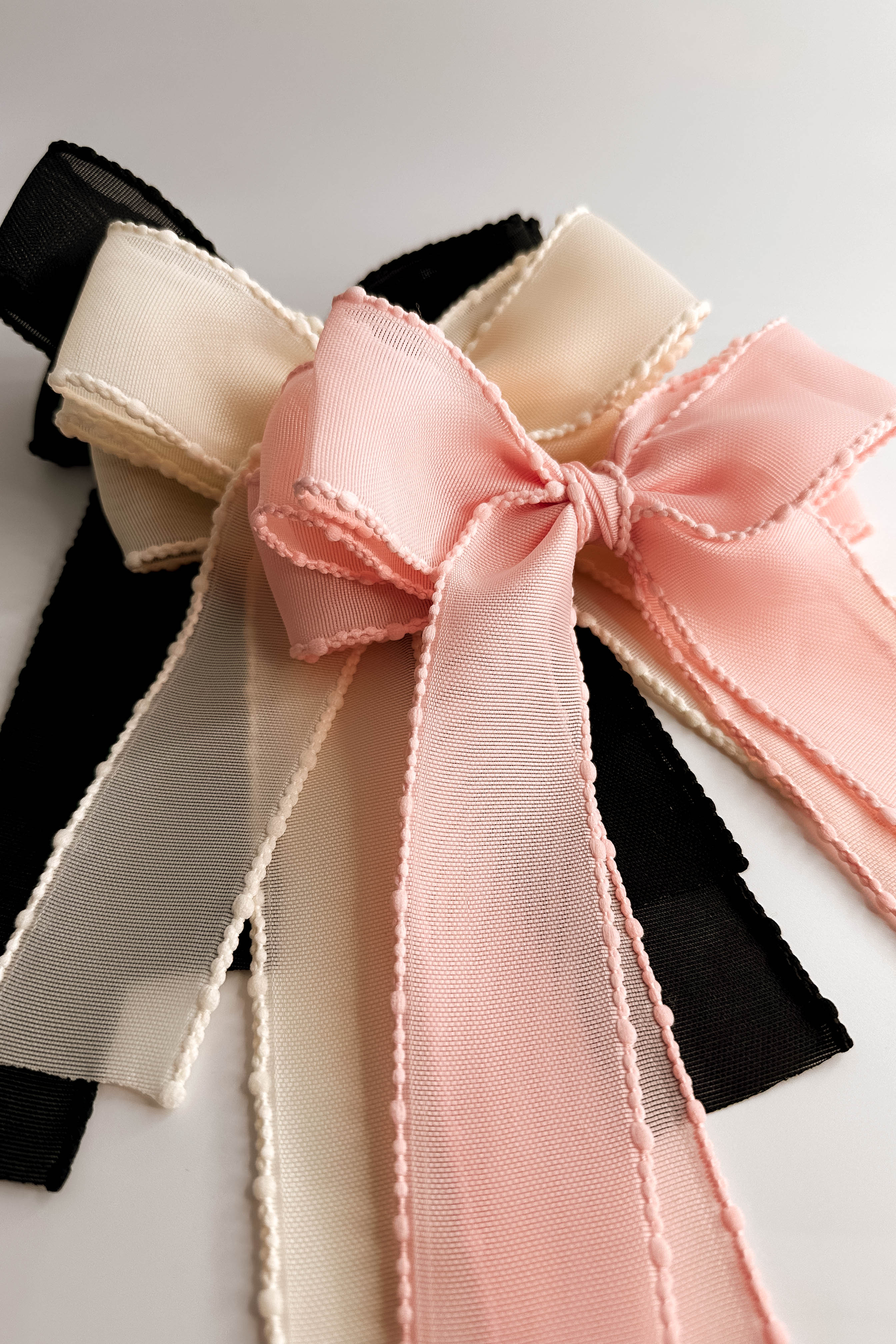 Close up view of the Harper Sheer Hair Bow Barrette in Black, Ivory, and Pink layered one each other against white background.
