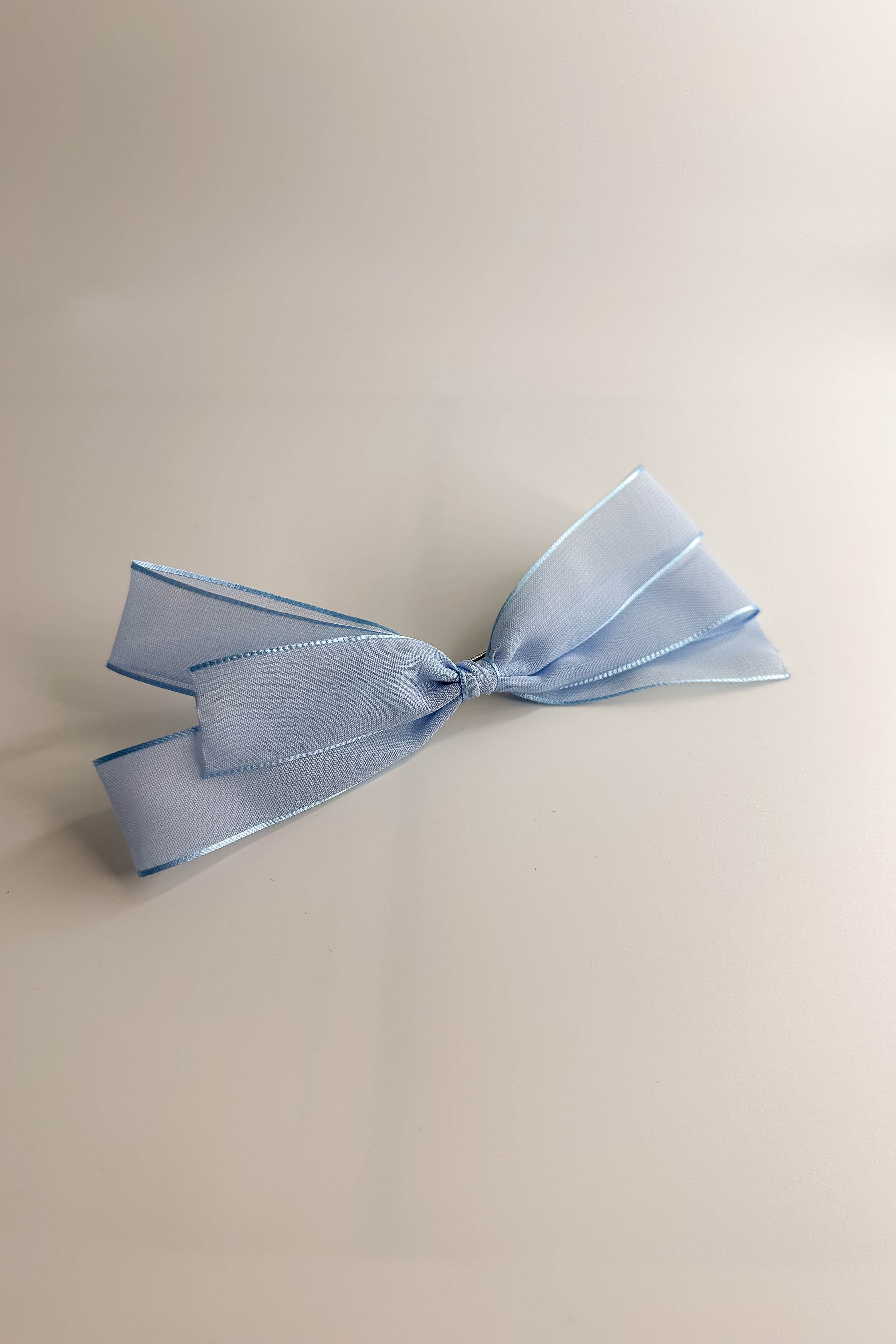 Flat lay view of the Amelia Sheer Hair Bow Barrette in Light Blue which features light blue sheer fabric, light blue satin trim, double bow with a french barrette clip.