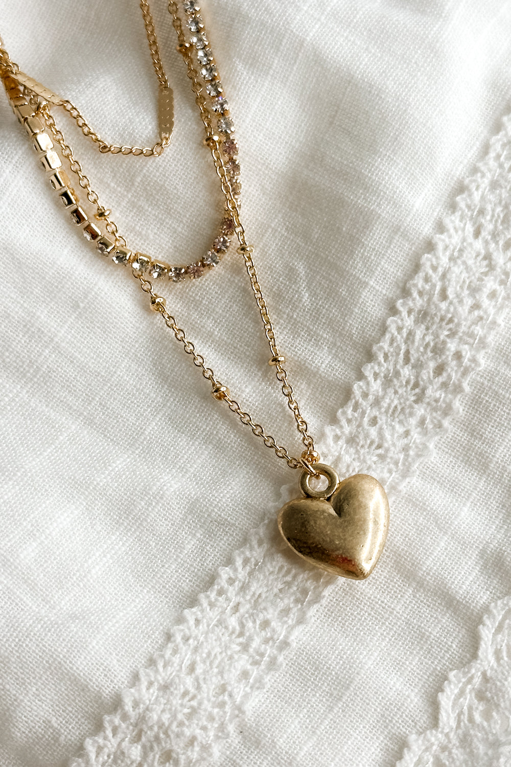 Close up view of the Celeste Gold Triple Layer Chain Heart Necklace which features two gold chain link layers, rhinestone layer, gold heart medallion and set in golds