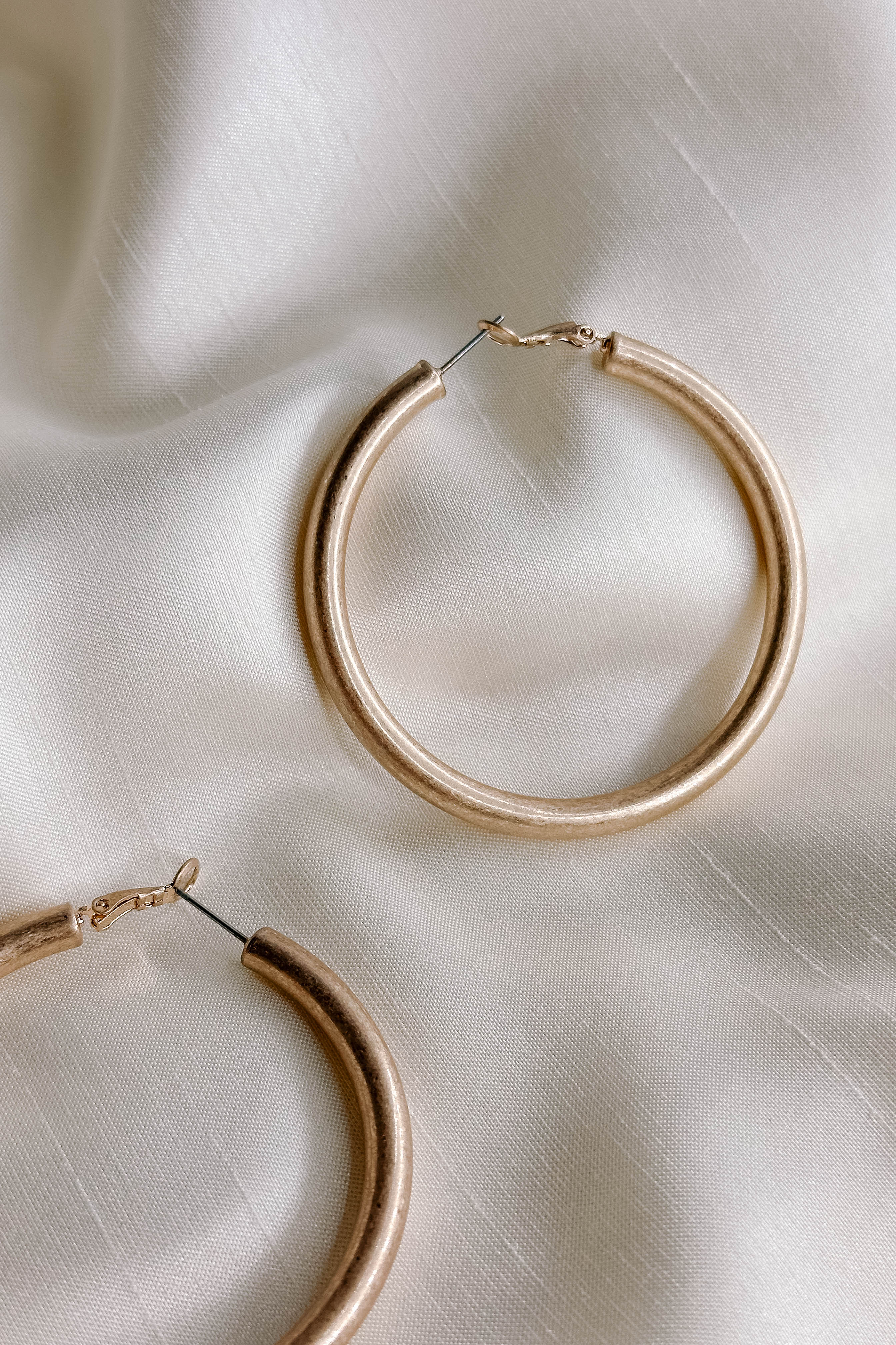 Close up single view of the Lucy Gold Brushed Hoop Earrings that features closed, medium brushed gold hoops with a hinged closure, shown on cream fabric.