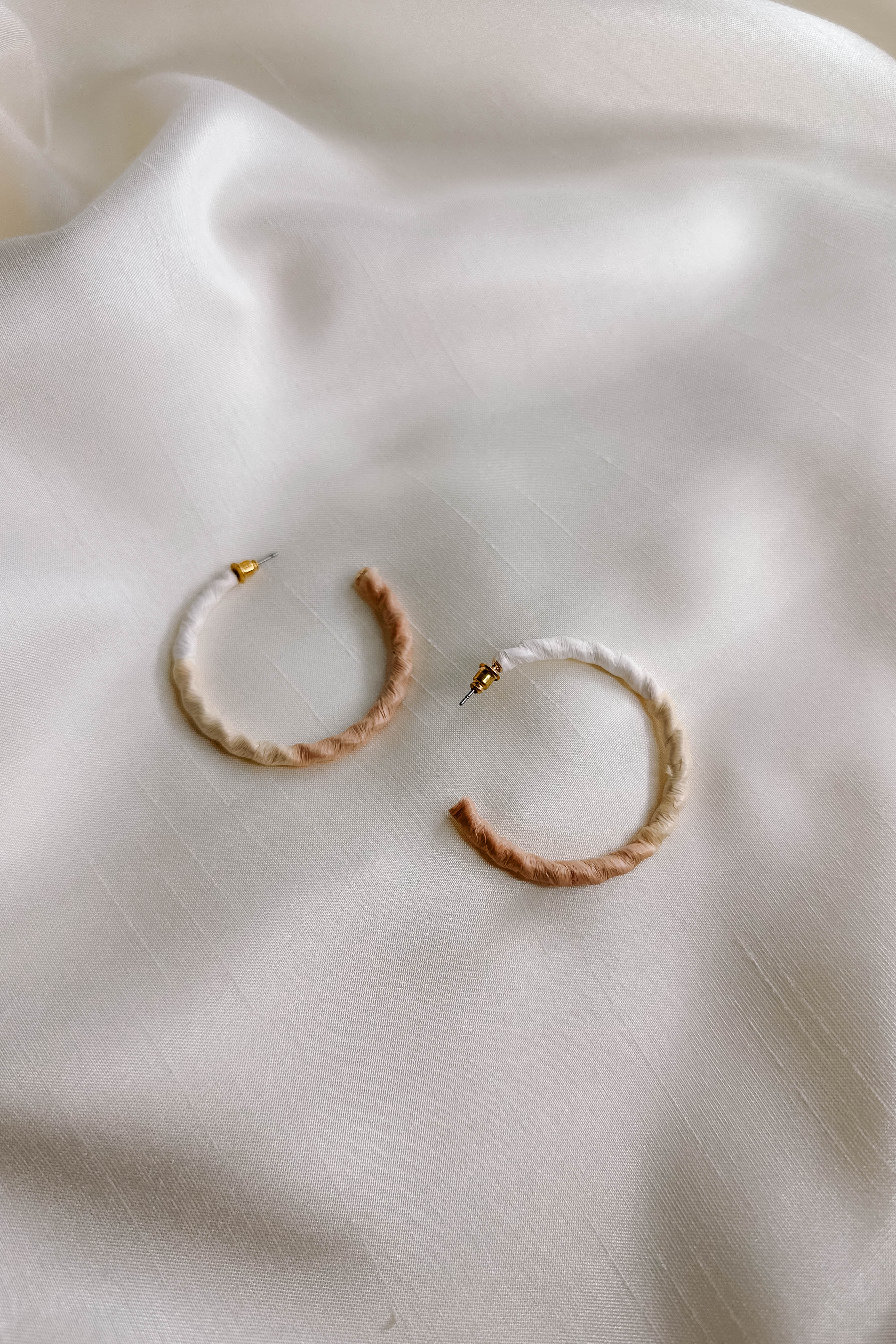 Side view of the Summer Breeze Earrings which features open, large hoops wrapped in white, beige and cream paper, shown on cream fabric.