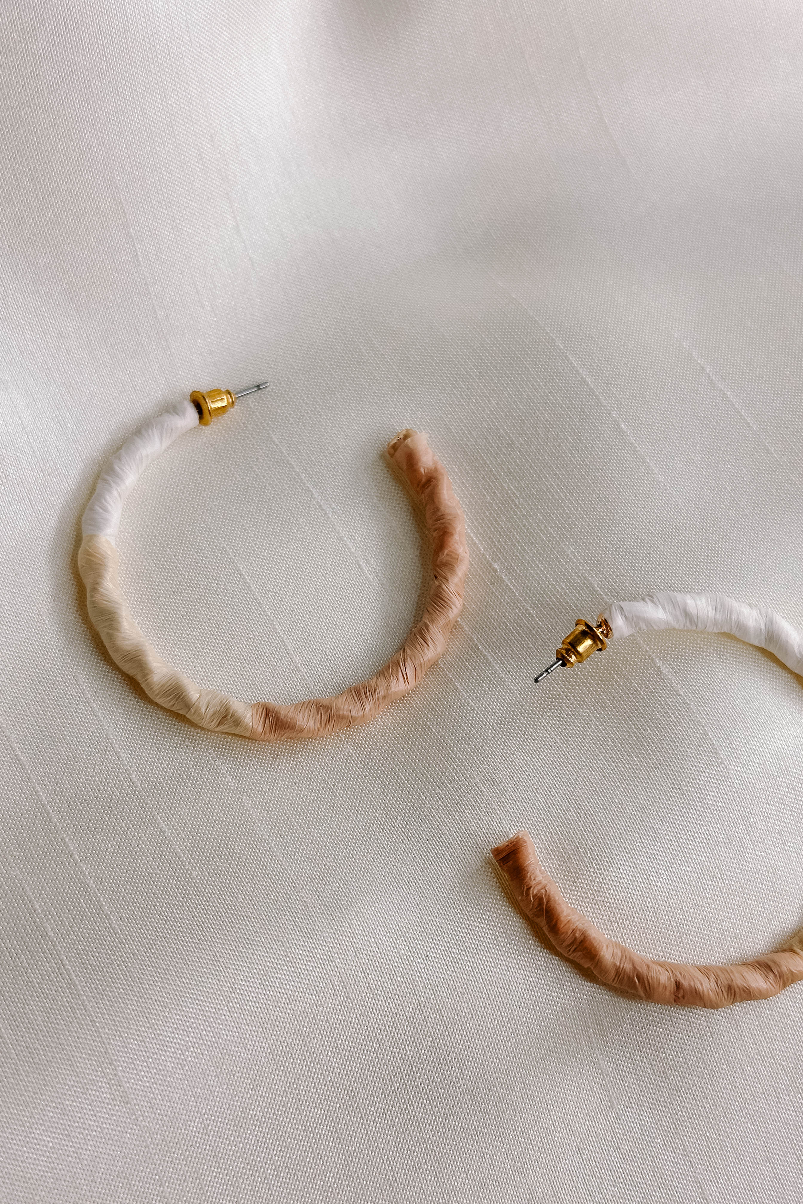 Side view of the Summer Breeze Earrings which features open, large hoops wrapped in white, beige and cream paper, shown on cream fabric.