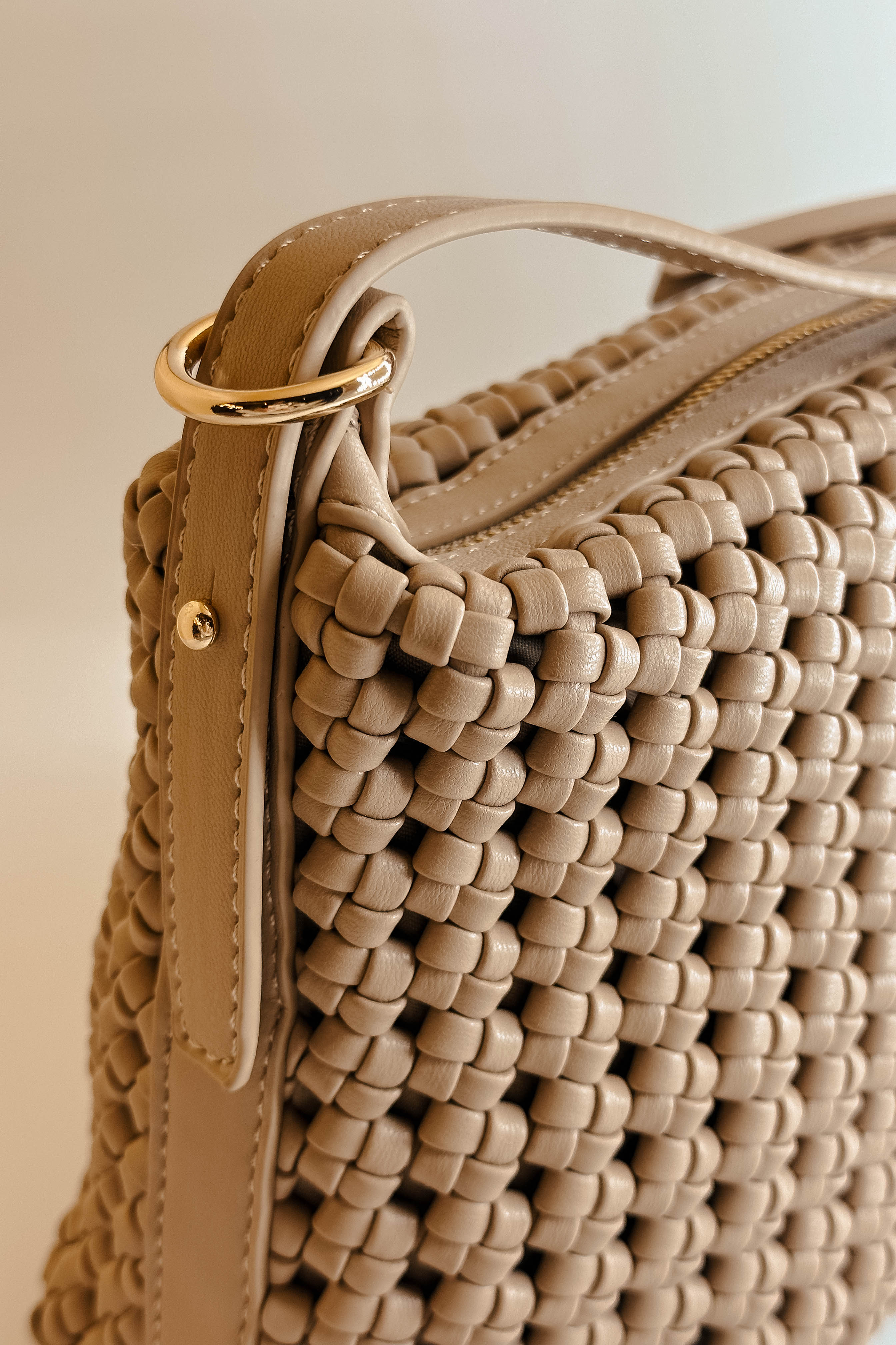 Close detailed side view of the Naya Beige Woven Purse.  It has dark beige faux-leather material with a woven design, short and long removable straps, gold hardware, and a patterned interior with a zippered pocket and a slit pocket.