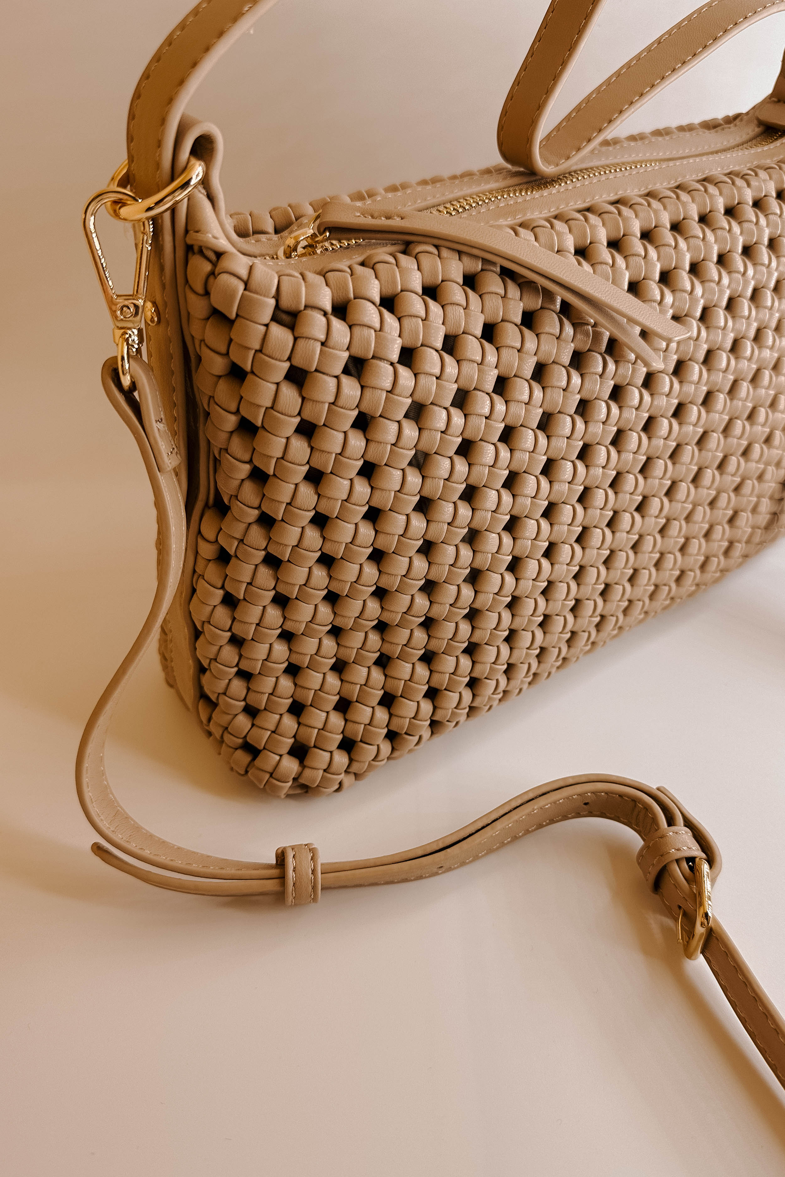 Close front view of the Naya Beige Woven Purse. It has dark beige faux-leather material with a woven design, short and long removable straps, gold hardware, and a patterned interior with a zippered pocket and a slit pocket.