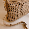 Close front view of the Naya Beige Woven Purse. It has dark beige faux-leather material with a woven design, short and long removable straps, gold hardware, and a patterned interior with a zippered pocket and a slit pocket.