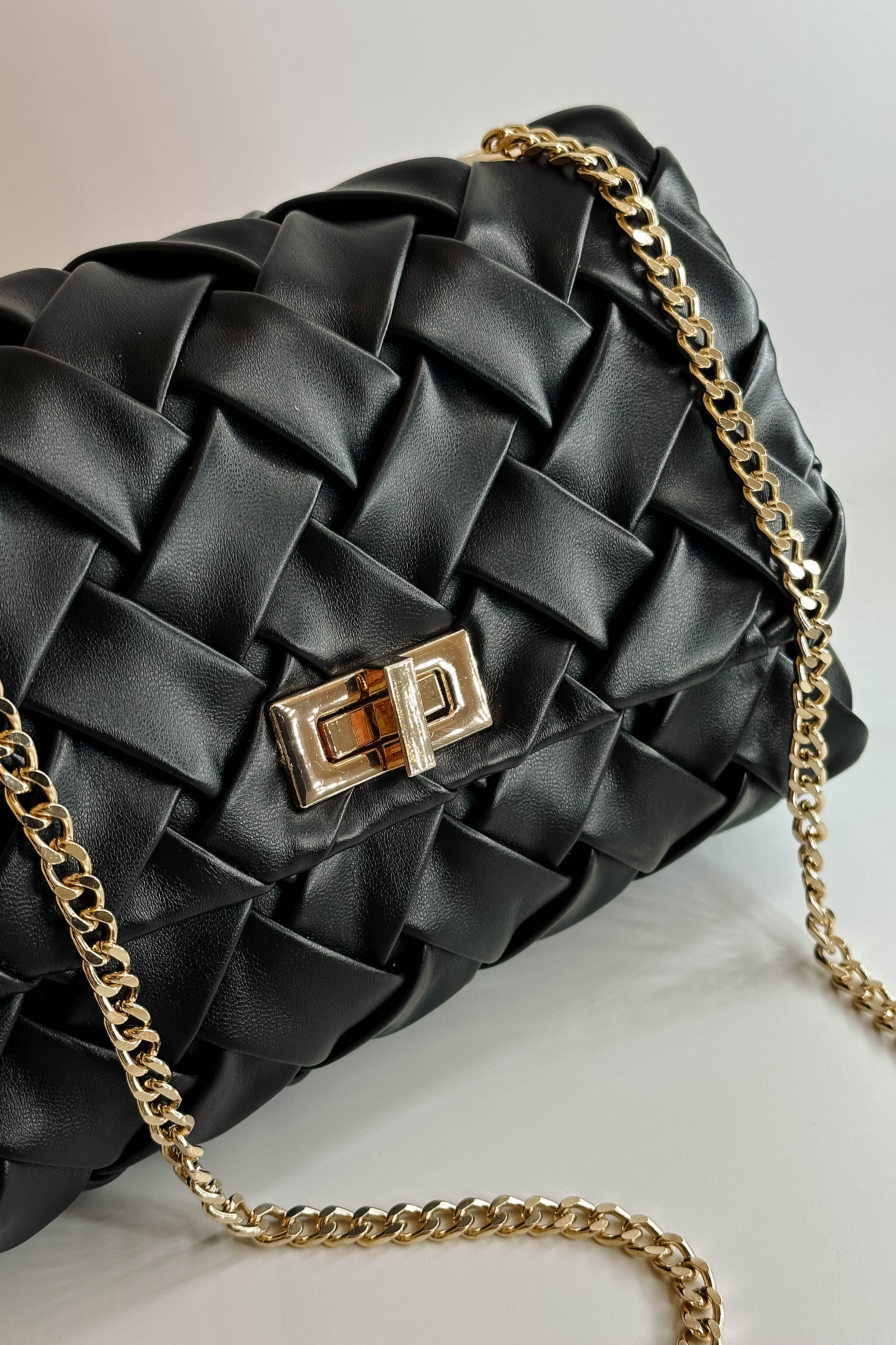 Close up view of the Carrie Black Leather Woven Purse which features black woven leather fabric, gold button closure and gold chain link strap.