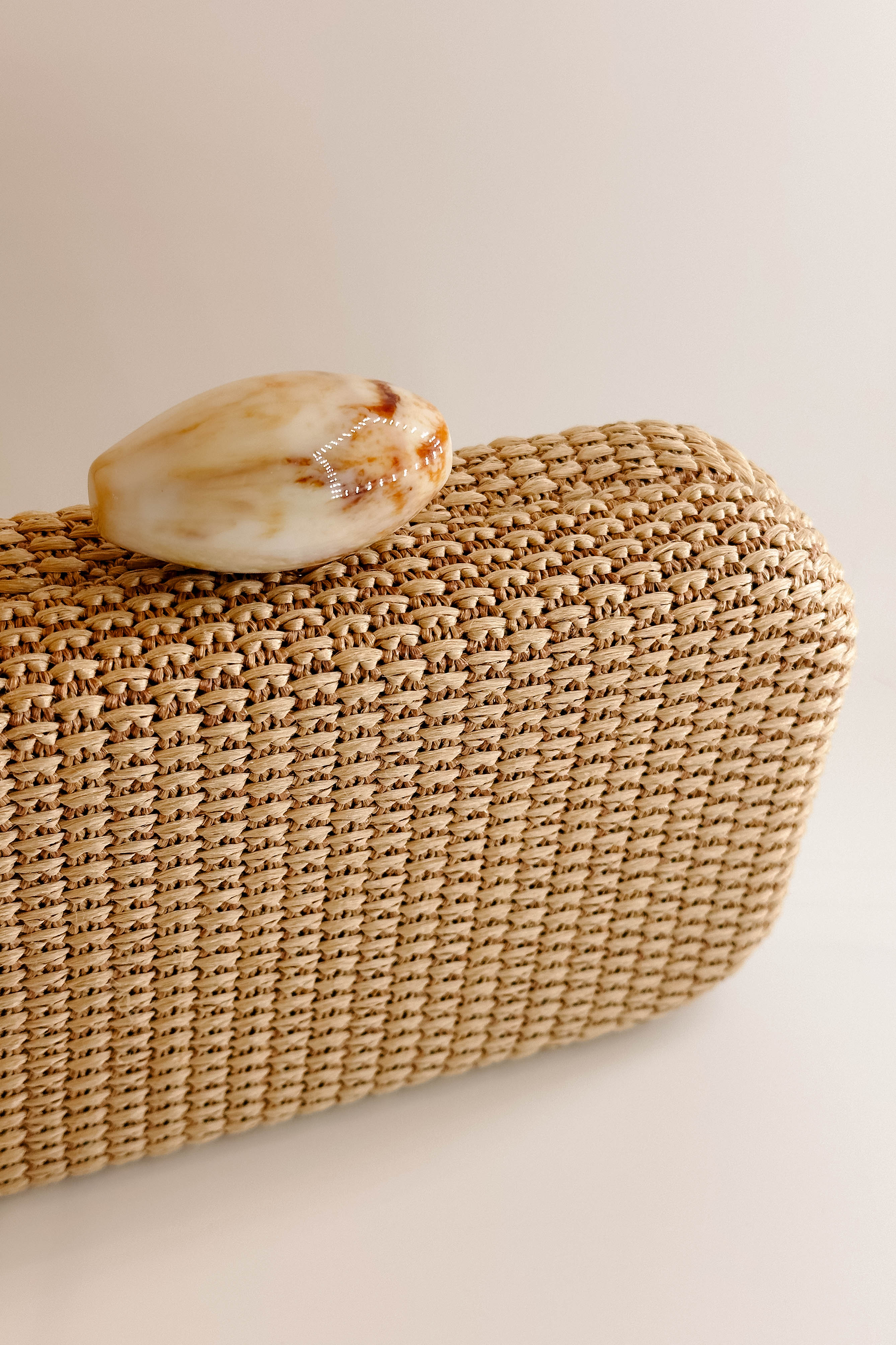 Detailed view of the pearl closure on the Frankie Clutch. It features a woven natural texture with an oval pearl closure. On the inside, is a gold chain attachment to be transformed as a crossbody.