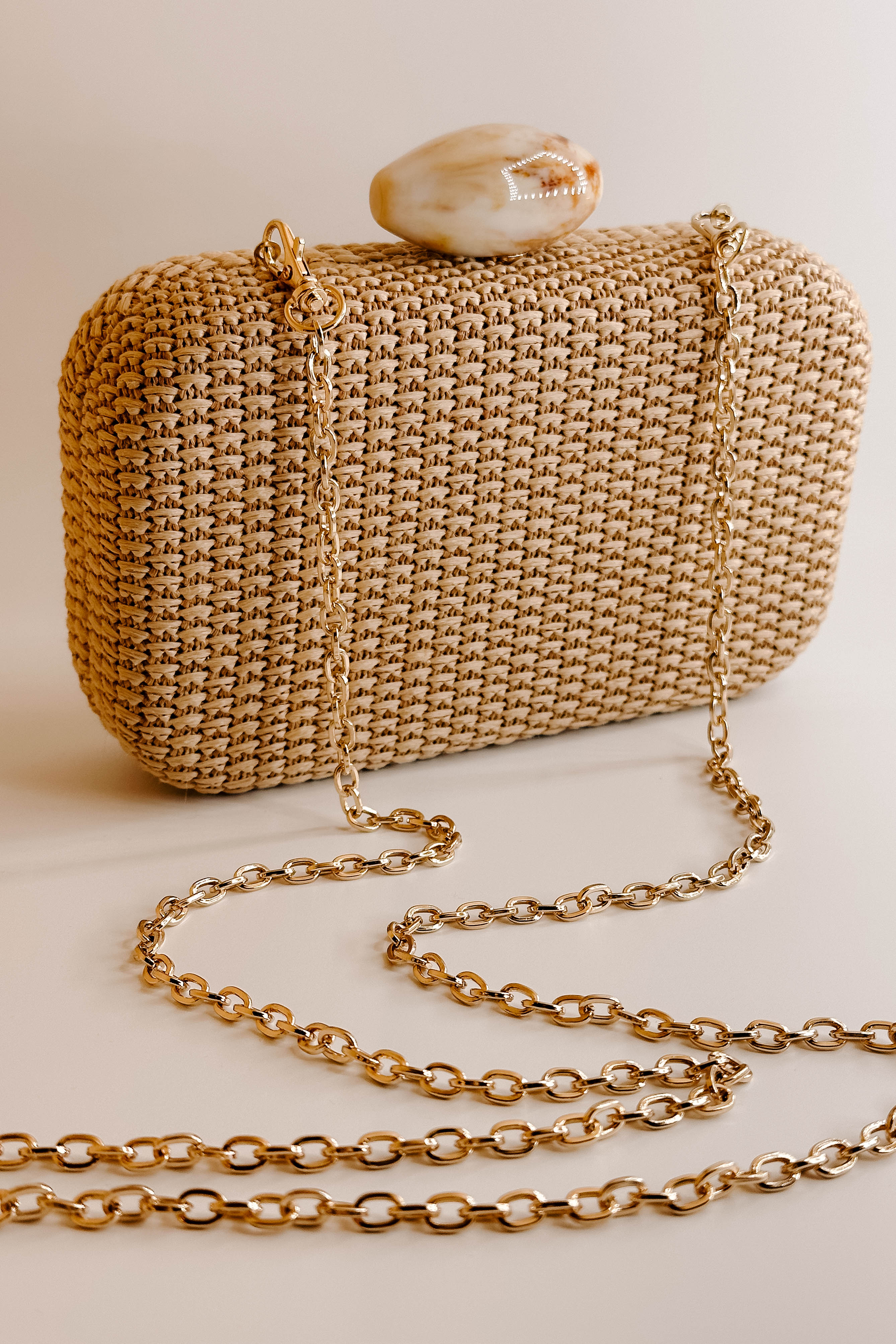 Detailed view of the Frankie Clutch. It features a woven natural texture with an oval pearl closure. On the inside, is a gold chain attachment to be transformed as a crossbody.