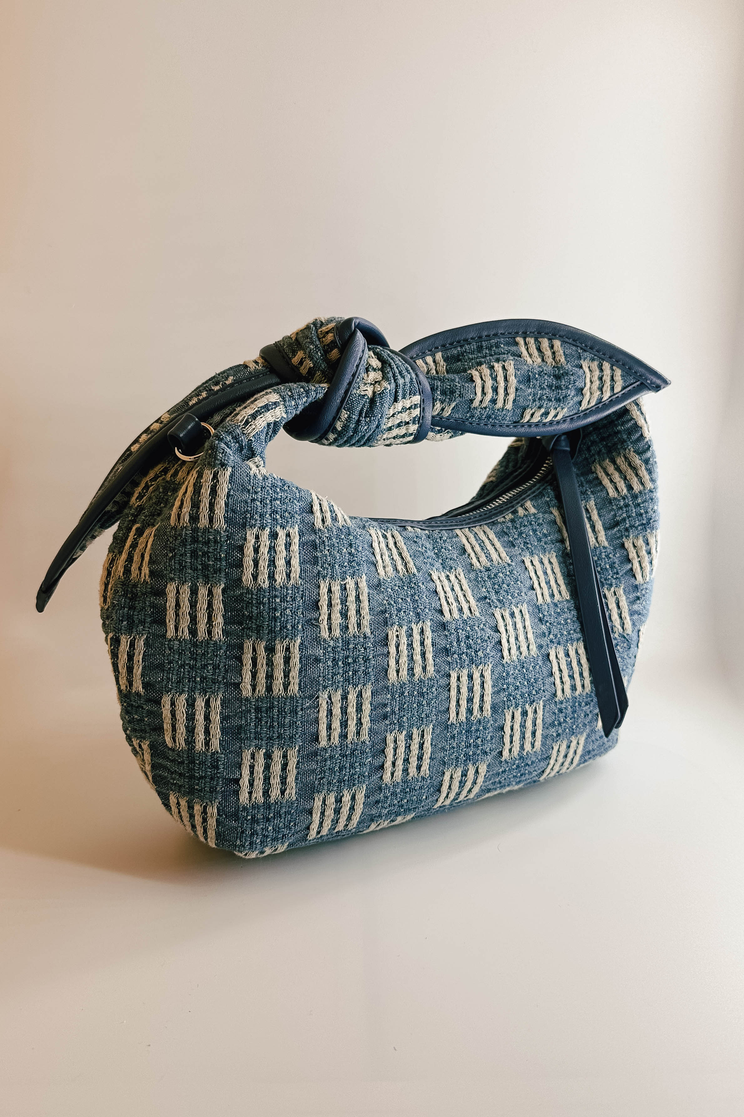 A close up front view of The Miley Denim Knot Handbag. This features a medium wash denim and tan pattern with a knot handle. There is a zipper closure along with a pocket on the inside.