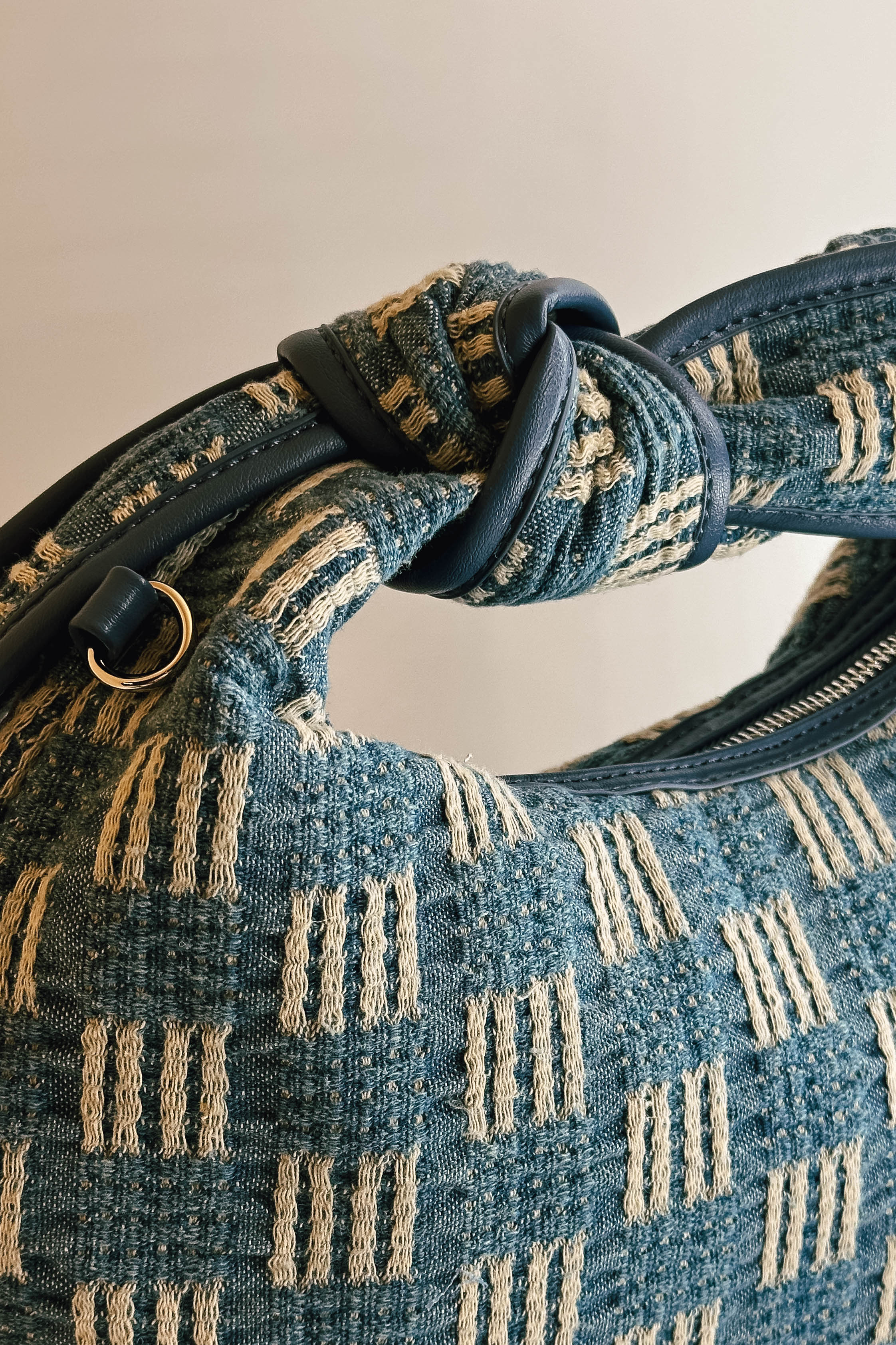 A close up handle view of The Miley Denim Knot Handbag. This features a medium wash denim and tan pattern with a knot handle. There is a zipper closure along with a pocket on the inside.