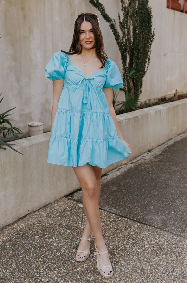 Full body view of female model wearing the  Gloria Light Blue Short Sleeve Mini Dress which features aqua blue cotton fabric, aqua blue lining, mini length, ruffle tiered details, a sweetheart neckline with drawstring ties, a smocked back, and short puff sleeves.