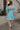 Full body back view of female model wearing the Gloria Light Blue Short Sleeve Mini Dress which features aqua blue cotton fabric, aqua blue lining, mini length, ruffle tiered details, a sweetheart neckline with drawstring ties, a smocked back, and short puff sleeves.