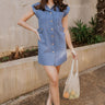 Full body view of female model wearing the Rebecca Blue Denim Sleeveless Dress which features blue denim fabric, a button-up front with brown wooden buttons, a collared neckline, chest pockets, side pockets, and a sleeveless design. 
