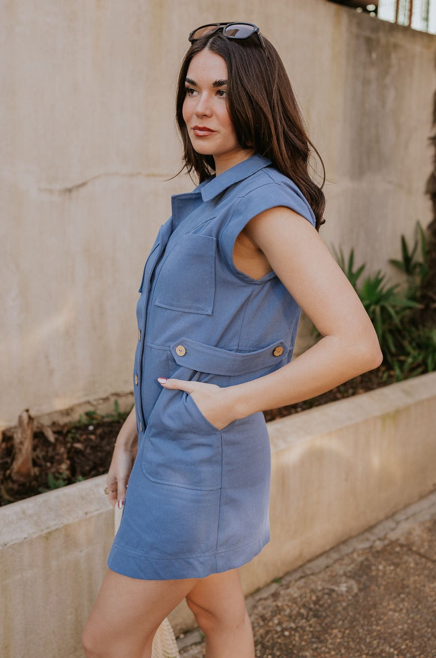 Side view of female model wearing the Rebecca Blue Denim Sleeveless Dress which features blue denim fabric, a button-up front with brown wooden buttons, a collared neckline, chest pockets, side pockets, and a sleeveless design.