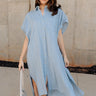 Full body front view of model wearing the Selah Chambray Button Up Midi Dress that has light blue cotton fabric with monochrome plaid print, midi length, slits on each side, a left front chest pocket, light cream tortoise button closures, a collared neckline, and short sleeves.