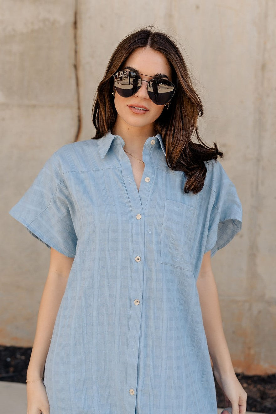 Upper front view of model wearing the Selah Chambray Button Up Midi Dress that has light blue cotton fabric with monochrome plaid print, midi length, slits on each side, a left front chest pocket, light cream tortoise button closures, a collared neckline, and short sleeves.