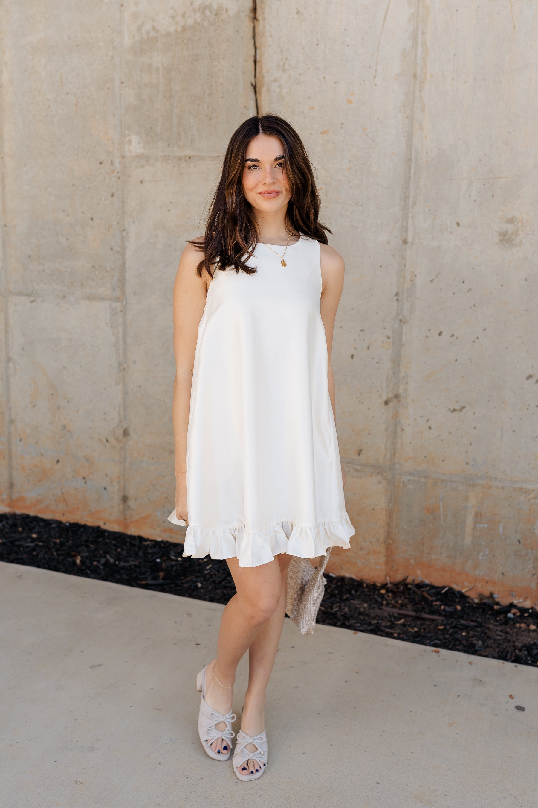 front image of model wearing the Anastasia Ruffle Mini Dress features cream sheen fabric, mini length, ruffle flare hem, cream lining, pockets on each side, round neckline, sleeveless and pearl button down closure on the back.