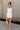 Full body front view of model wearing the Brielle White Sleeveless Mini Dress that features white stretchy fabric, mini length, white lining, a layered skirt, pleated cinched side detail, a square neckline, adjustable straps, and monochrome back zipper with a hook closure.