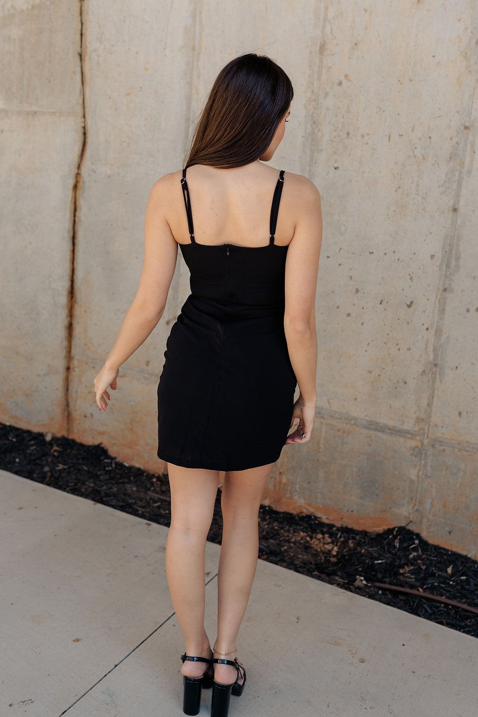 Full body back view of the model wearing the Elodie Black Rhinestone Bow Mini Dress that has black knit fabric, black lining, mini length, scooped neckline, adjustable straps, removable rhinestone bows, sleeveless and monochrome back zipper with hook closure.