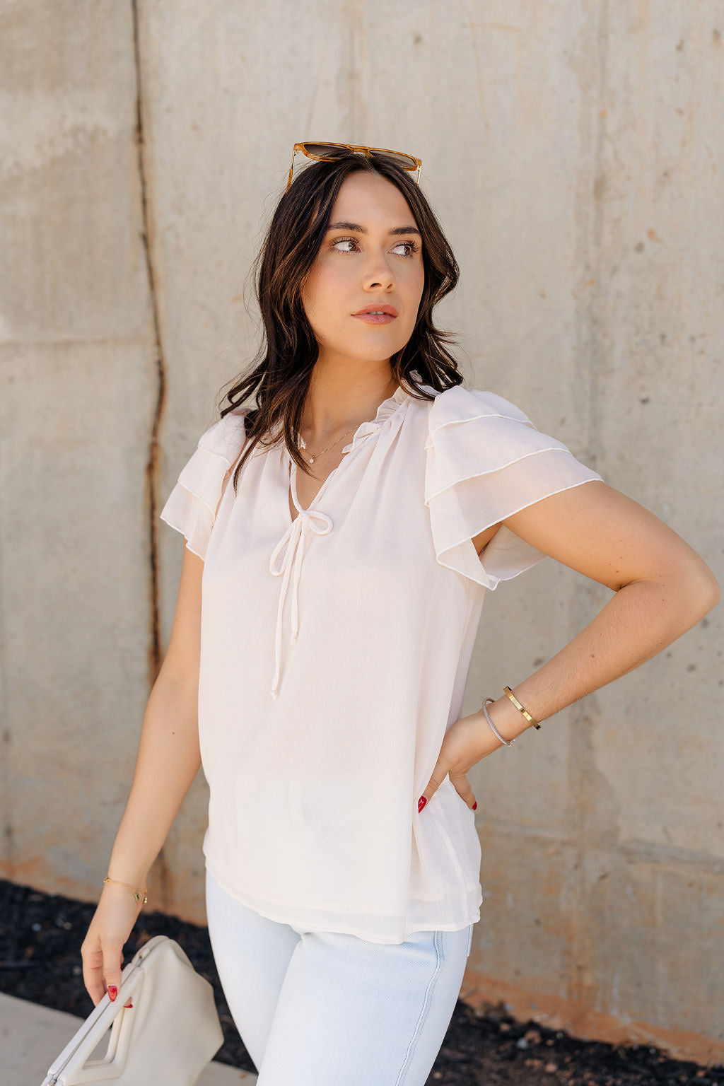 Side view of model wearing the Olivia Cream Ruffle Short Sleeve Top which features cream sheer fabic, cream lining, ruffle neckline with drawstring ties, and ruffle tiered short sleeves.