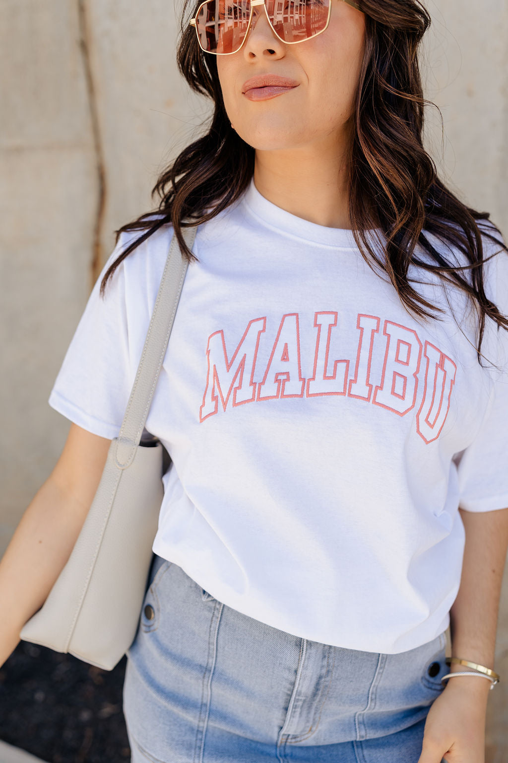 Close up view of model wearing the Malibu White & Blush Short Sleeve Graphic Top which features white knit fabic, round neckline, shirt sleeves. Graphic says "MALIBU" in block letters with blush pink stitch.