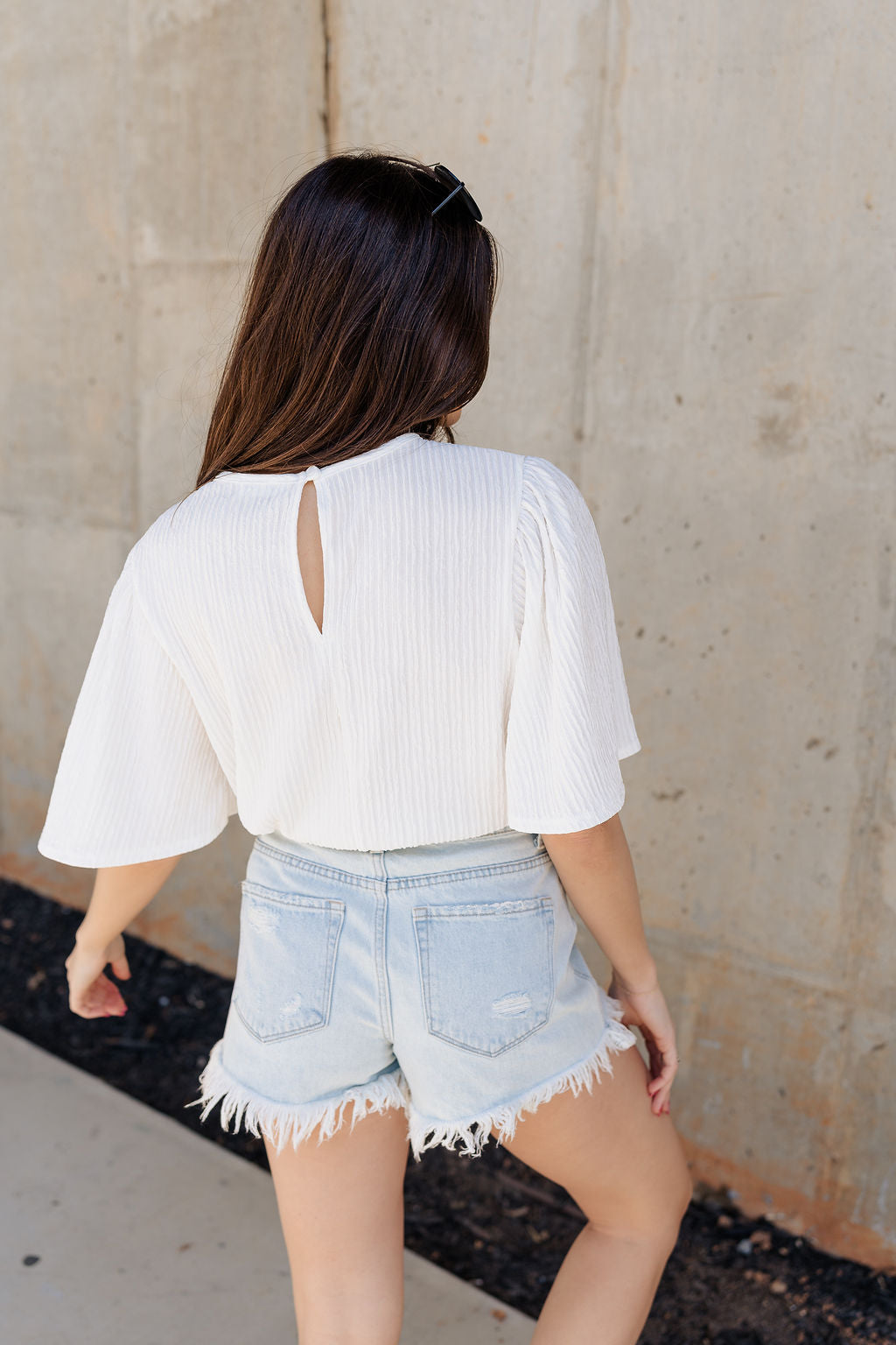 Back view of model wearing the Merritt Ivory Short Sleeve Top which features ivory textured fabric, round neckline, short sleeves and back key hole with monochrome button closure.