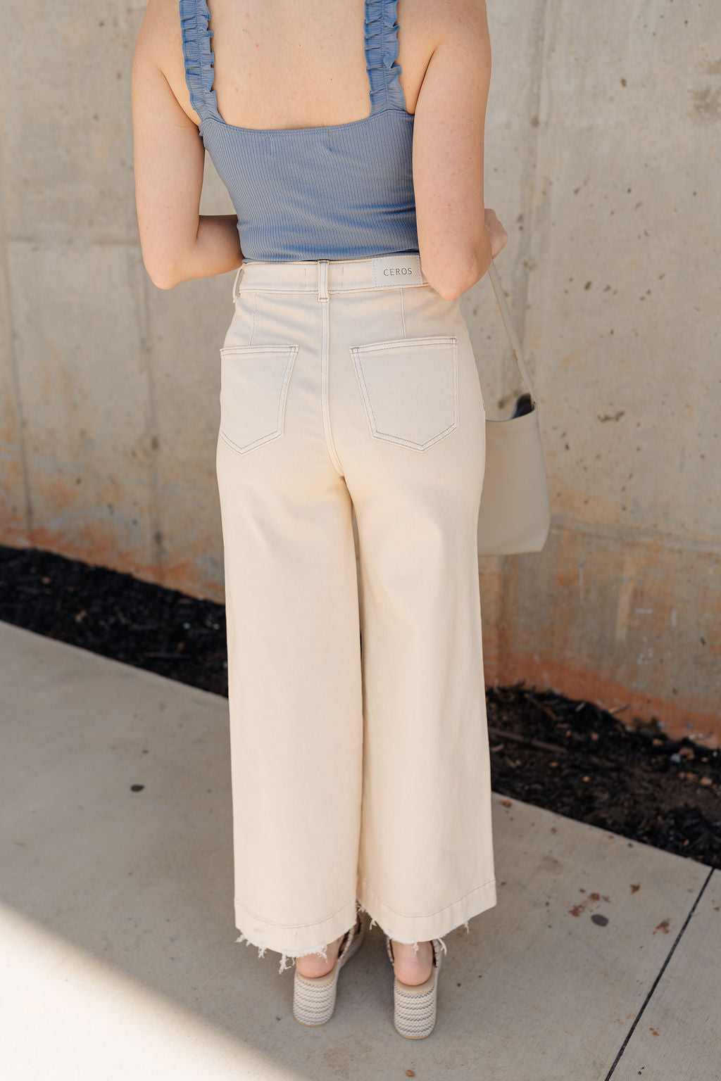 Back view of model wearing the Ceros: Florence Ecru Wide Leg Jeans which features ecru denim fabric, a front zipper with a button closure, belt loops, a super high-rise waist, two front pockets, two back pockets, and wide cropped legs with distressed hems.