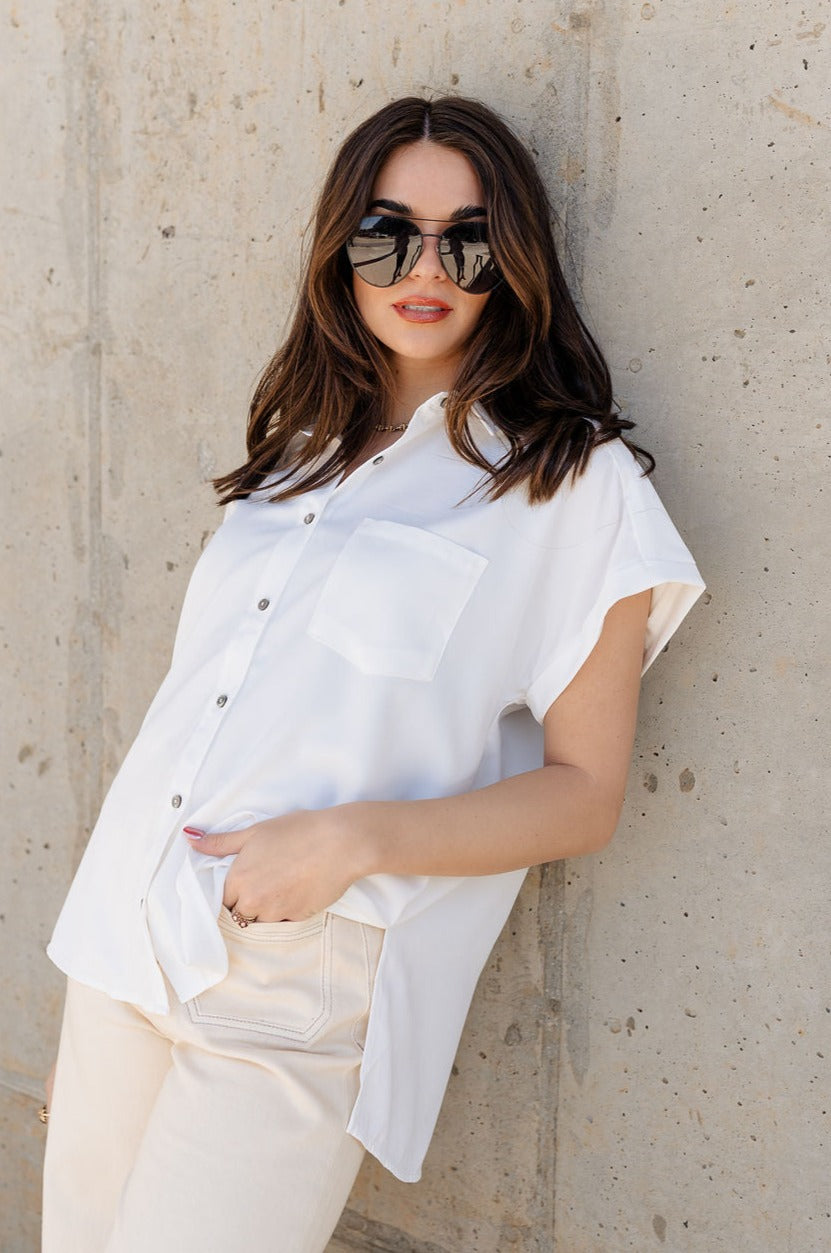Front view of model wearing the Claire Off White Collared Short Sleeve Top which features white fabric, a front chest pocket, short sleeves with cuffs, small side slits, a button-up front, and a collared neckline.