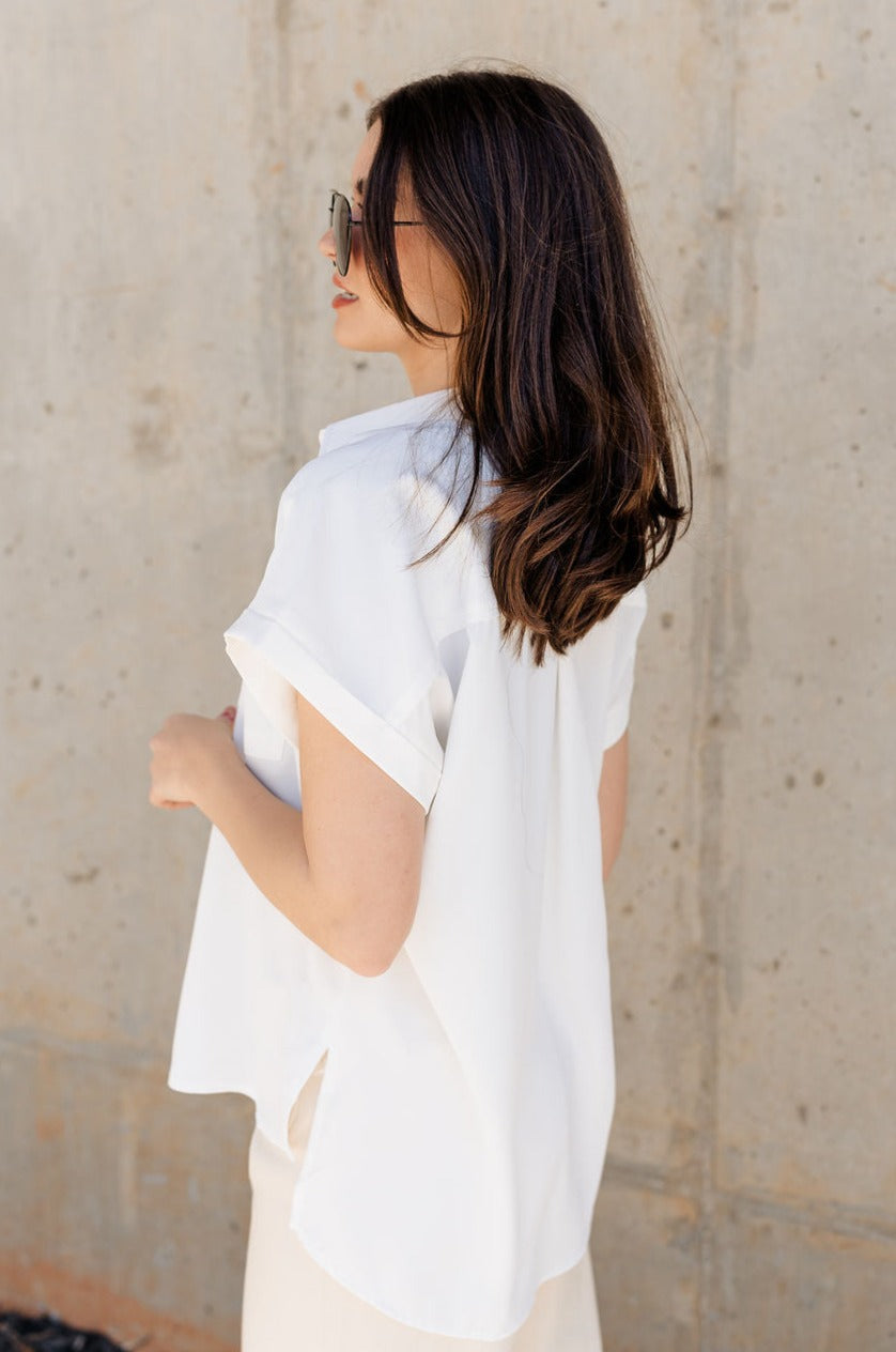 Side view of model wearing the Claire Off White Collared Short Sleeve Top which features white fabric, a front chest pocket, short sleeves with cuffs, small side slits, a button-up front, and a collared neckline.