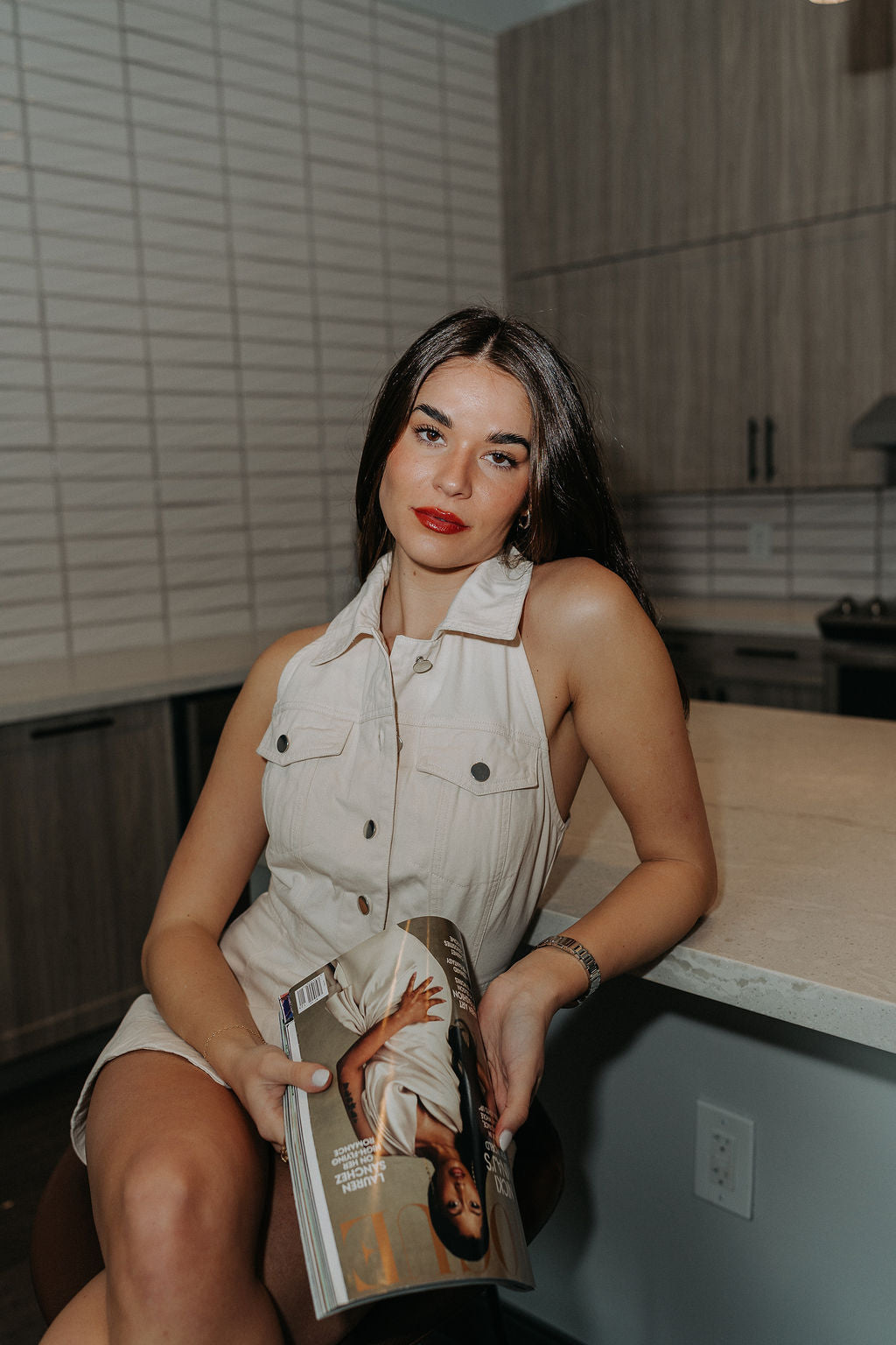 Model is sitting while wearing the cream colored Eleanor Sleeveless Button Front Mini Dress that has cotton fabric, a button-up front with silver buttons, buttoned chest pockets, a collared neckline, and a racerback cut.