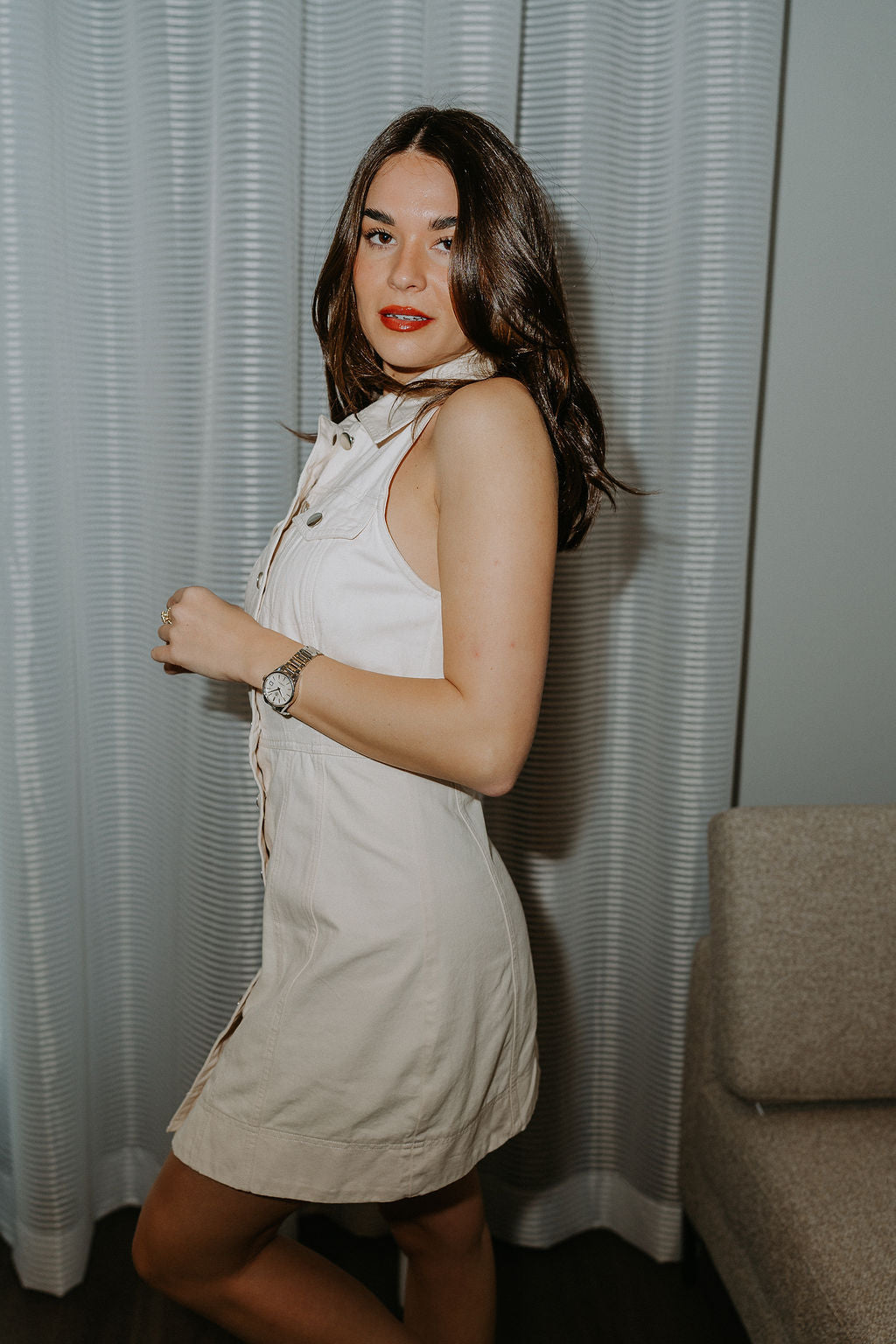 Close side view of model wearing the cream colored Eleanor Sleeveless Button Front Mini Dress that has cotton fabric, a button-up front with silver buttons, buttoned chest pockets, a collared neckline, and a racerback cut.