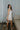 Back view of model wearing the cream colored Eleanor Sleeveless Button Front Mini Dress that has cotton fabric, a button-up front with silver buttons, buttoned chest pockets, a collared neckline, and a racerback cut.
