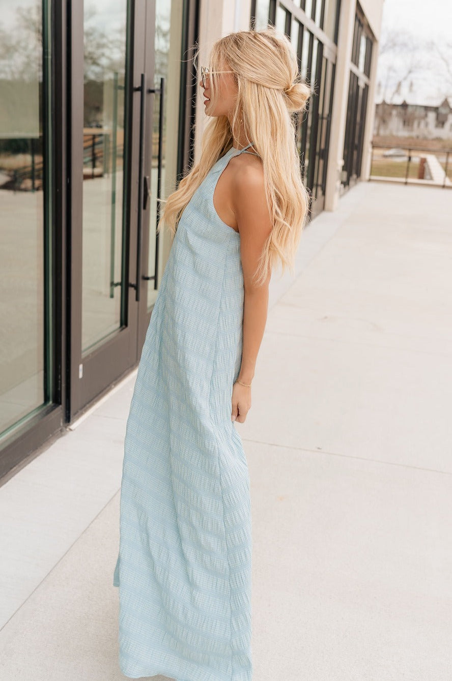 Full body side view of model wearing the Gillie Light Blue Halter Maxi Dress which features light blue and yellow knit textured fabric, light blue lining, small gingham print design, maxi length, halter neckline, adjustable straps, sleeveless and open back.