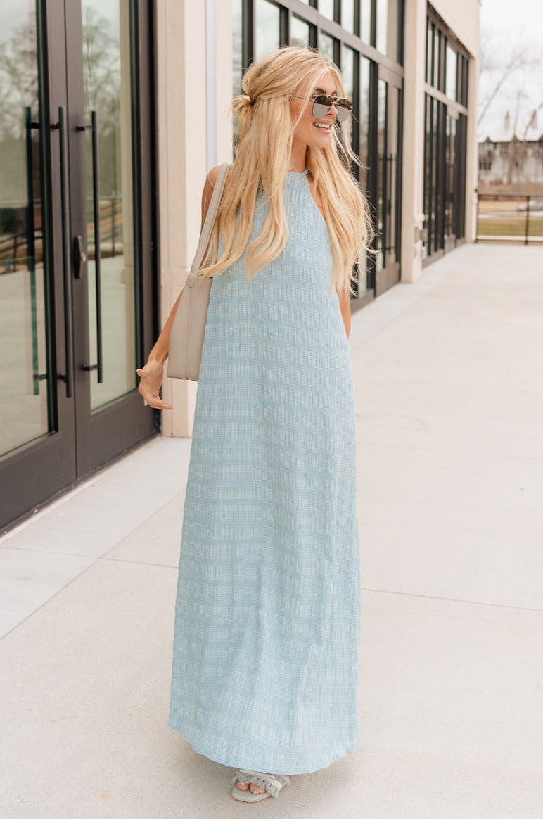 Full body view of model wearing the Gillie Light Blue Halter Maxi Dress which features light blue and yellow knit textured fabric, light blue lining, small gingham print design, maxi length, halter neckline, adjustable straps, sleeveless and open back.