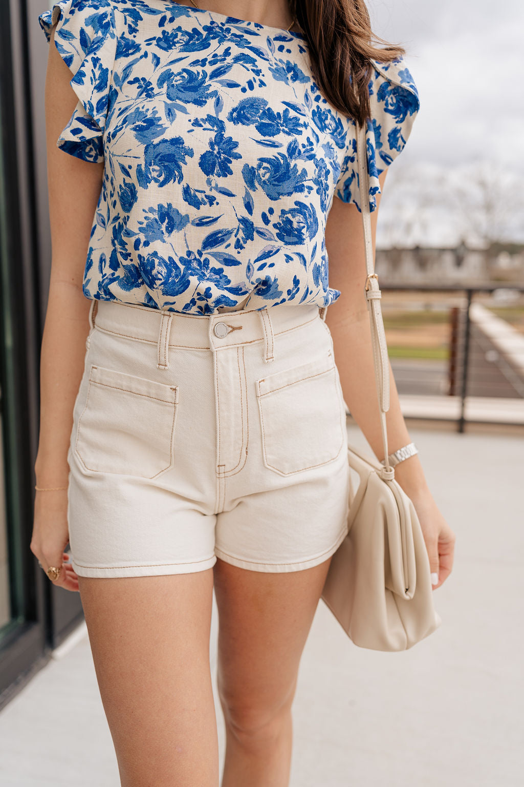 Close-up front view of female brunette model wearing the Mckenzie Off White Denim Shorts that feature off white denim, contrast stitching, pockets, and a high rise. Styled with blue and white floral top.