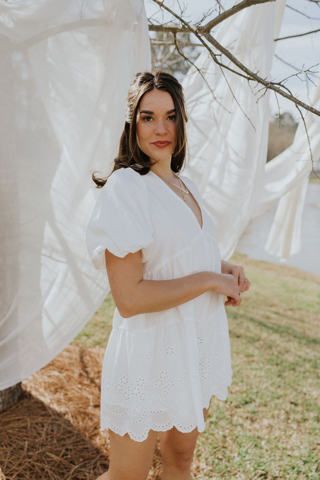 Frontal side view of female model wearing the Madeline White Eyelet Romper that has white fabric with eyelet details, short puff sleeves, a vneck, and a keyhole back with a tie.