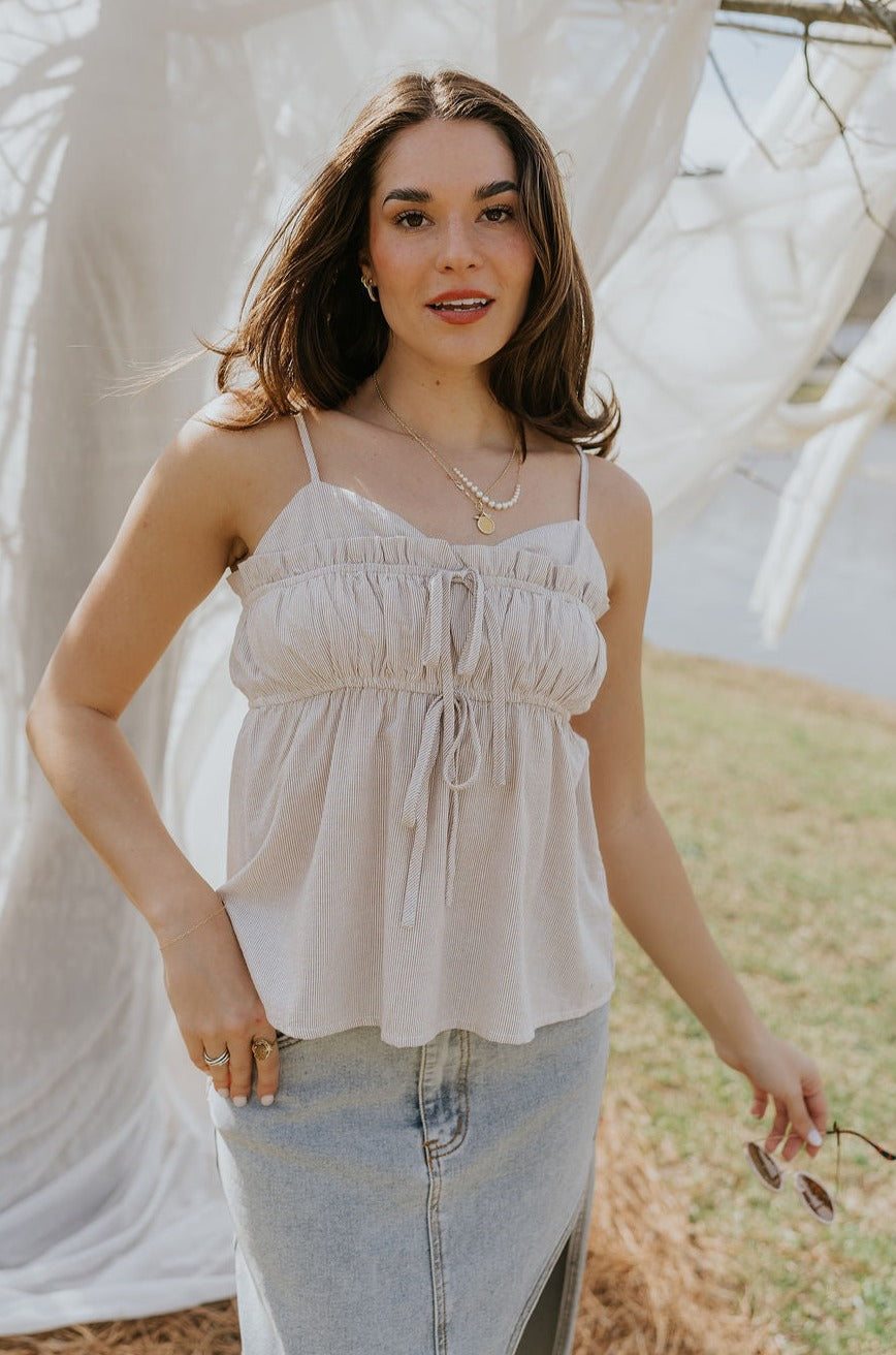 Front view of female model wearing the Billie White & Taupe Striped Tank that has thin vertical stripes, front tie details, ruffle trim, and spaghetti straps