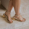 Right side view of female model wearing the Banita Sandal in Light Gold Crackled which features light gold crackled leather fabric, skinny criss-cross straps, square toe, slim ankle adjustable buckle and textured rattan heel